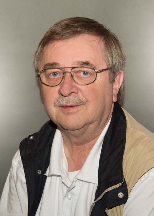 Portraitfoto eines lächelnden Mannes mit Brille und grauem Oberlippenbart. 