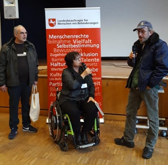 Vor der Bühne stehen Herr Kosciol, Frau Wontorra (im Rollstuhl) sowie Herr Altmann mit Mikrofonen in der Hand und begrüßen die Anwesenden. 